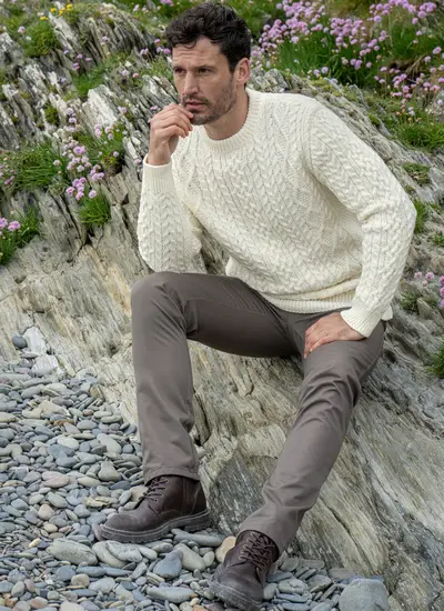 Man Sitting on Beachy Rock Wearing Oisin Aran Crew Neck Sweater in Natural